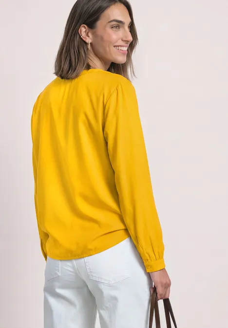Een vrouw in profiel draagt een mosterdgele blouse met lange mouwen, witte broek en houdt een tas vast.