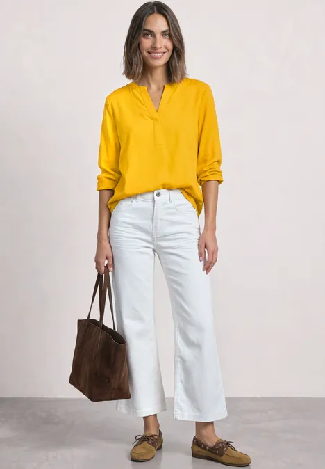 Vrouw in gele blouse, witte wijde jeans, bruine loafers en suède tas voor een lichte achtergrond.