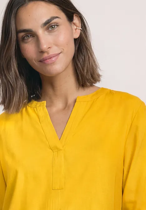 Portret van een glimlachende vrouw met halflang donker haar in een gele blouse tegen een lichte achtergrond.