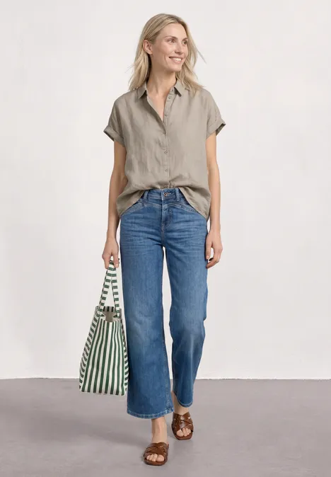 Dropped Shoulder Bluse aus Leinen timeless beige Frau in beigem Kurzarmhemd, blauen Jeans und braunen Sandalen mit grün-weißer Tasche.