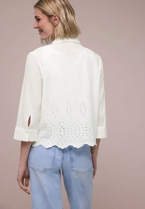 Rugaanzicht van een vrouw in een witte geborduurde blouse met driekwartmouwen en lichtblauwe jeans.