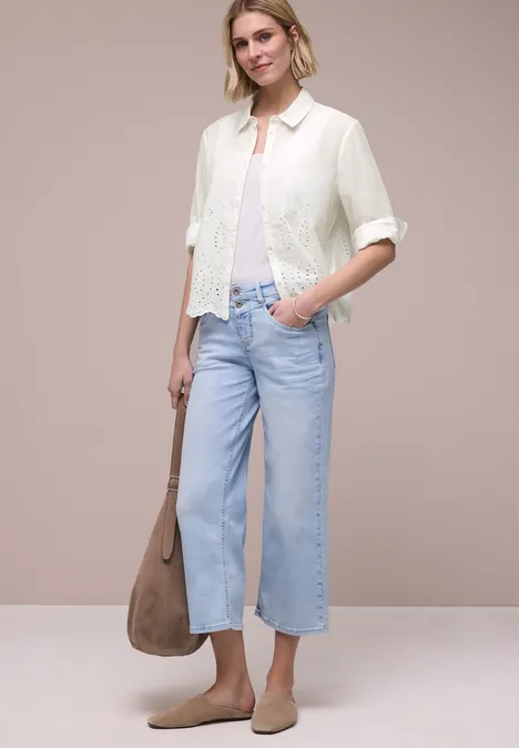 Vrouw in witte kanten blouse, lichtblauwe wijde cropped jeans, beige loafers en grote lichtbruine tas.