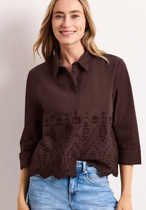 Vrouw draagt donkerbruine blouse met opengewerkte borduursels, geschulpte zoom en driekwartmouwen, gecombineerd met jeans.