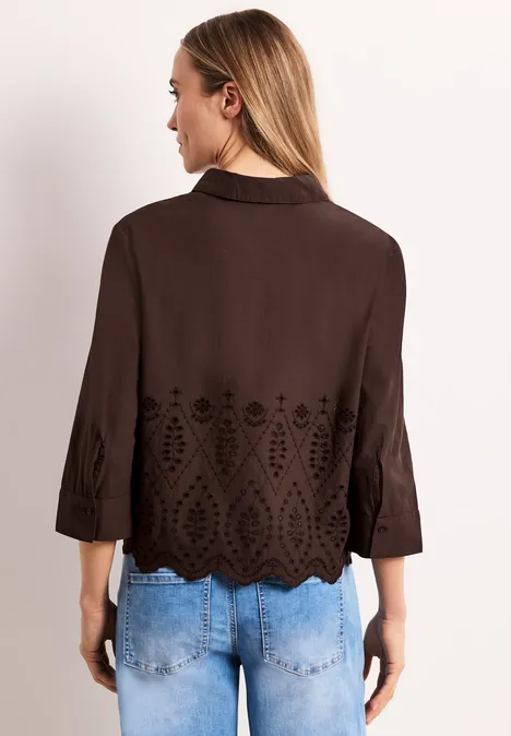 Vrouw van achteren in donkerbruine blouse met cutwork-geborduurde onderzoom en driekwartmouwen, bij lichte spijkerbroek.