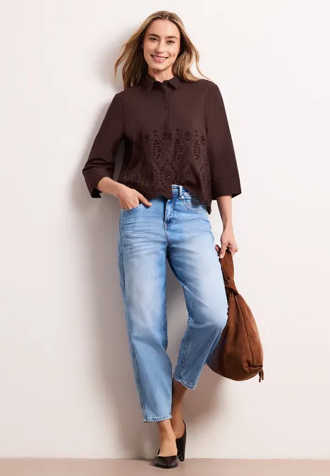 Vrouw met een bruine geborduurde blouse, lichtblauwe jeans, zwarte schoenen en bruine tas.