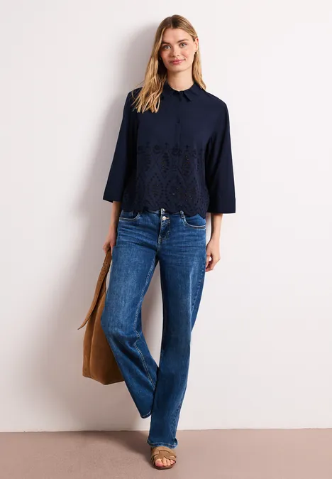 Vrouw in marineblauwe kanten top met driekwartmouwen, rechte jeans, sandalen en een bruine tas.