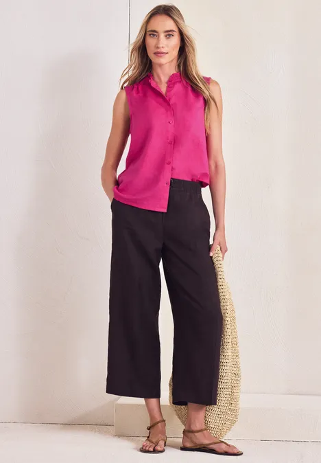 Frau in pinkem ärmellosem Top, schwarzen Culotte-Hosen, Sandalen und Strohtasche vor weißer Wand.