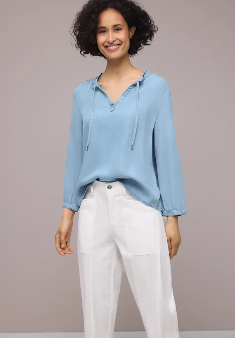 Vrouw in losse lichtblauwe blouse met capuchon en witte, wijde broek voor een neutrale achtergrond.
