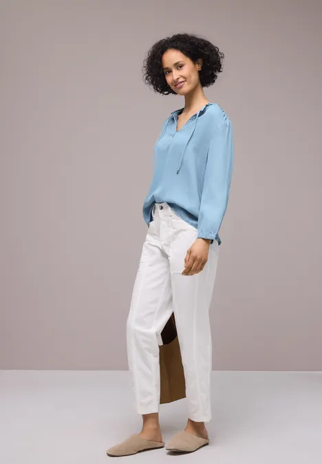 Vrouw in blauwe blouse, witte broek en bruine schoenen voor een effen achtergrond