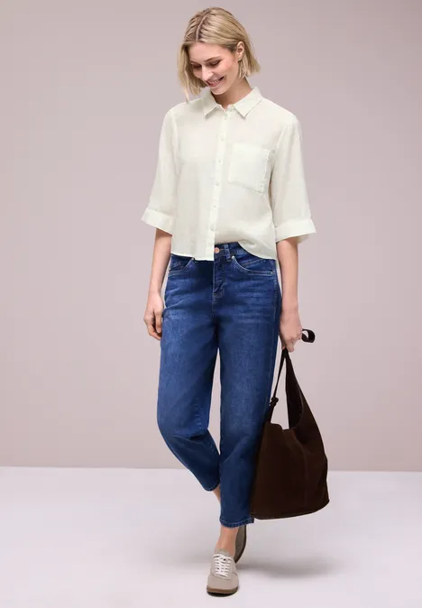 Femme en chemisier blanc et jean bleu, tenant un sac marron et des lunettes de soleil en main devant un fond neutre.