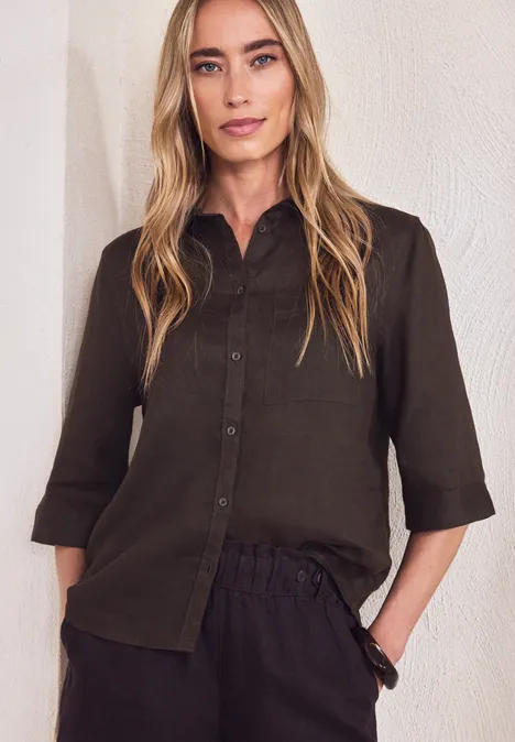 Personne en chemise olive à manches 3/4 et pantalon sombre devant un mur blanc.