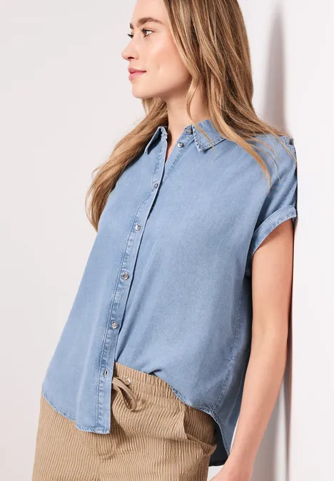 Een vrouw draagt een lichtblauw denimblouse met korte mouwen en een beige broek.
