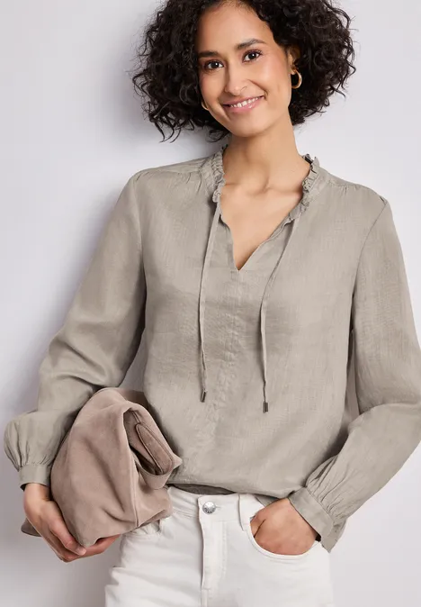 Vrouw in lichtbeige linnen blouse met ruches en koordsluiting, houdt bijpassend clutch, nonchalante look.