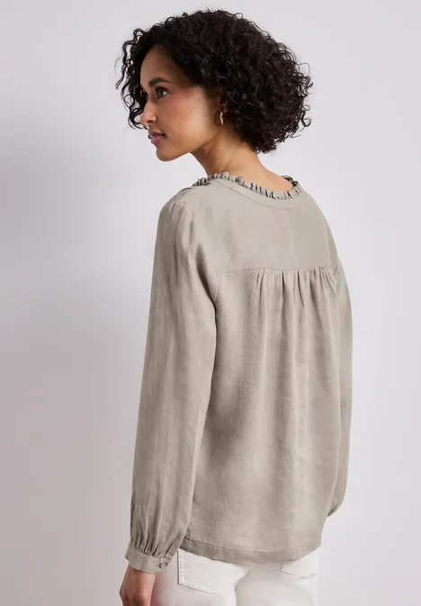 Vrouw van achteren in een losvallende grijze linnen blouse met plooien bij schouders en manchetten, lichte broek.