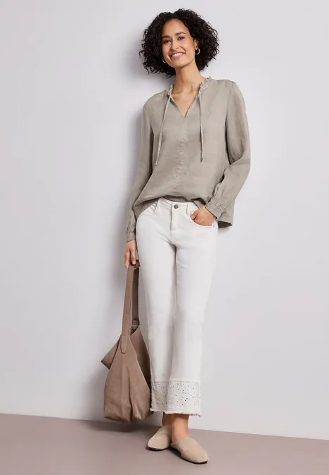 Persoon draagt lichtgrijs shirt, witte broek met kanten zoom, beige tas en platte schoenen tegen neutrale achtergrond.