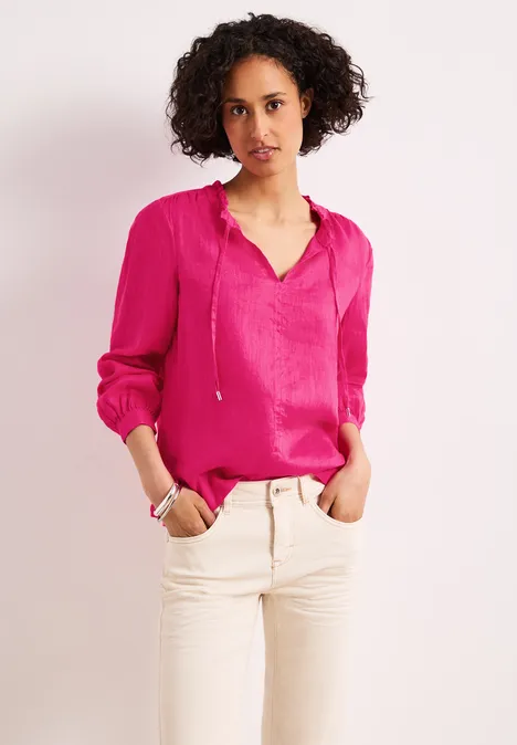 Vrouw in roze blouse met V-hals en lange mouwen, gecombineerd met een lichte broek voor een neutrale achtergrond.