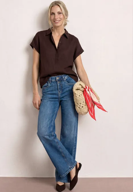 Vrouw in bruin kort shirt en blauwe jeans, met gevlochten rieten tas met rode sjaal en donker sleehakken.