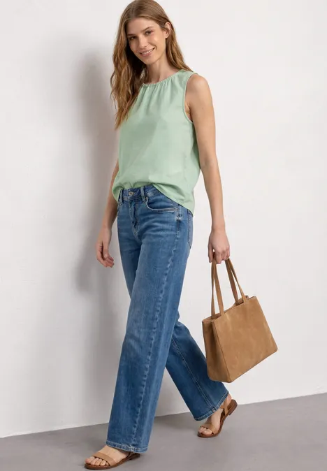 Frau trägt mintgrünes Top, weite Jeans, braune Sandalen und Handtasche vor weißer Wand.
