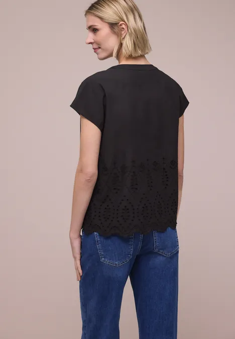 Vrouw van achteren in zwarte korte mouwenblouse met borduurwerk aan de zoom en blauwe spijkerbroek voor een beige achtergrond.