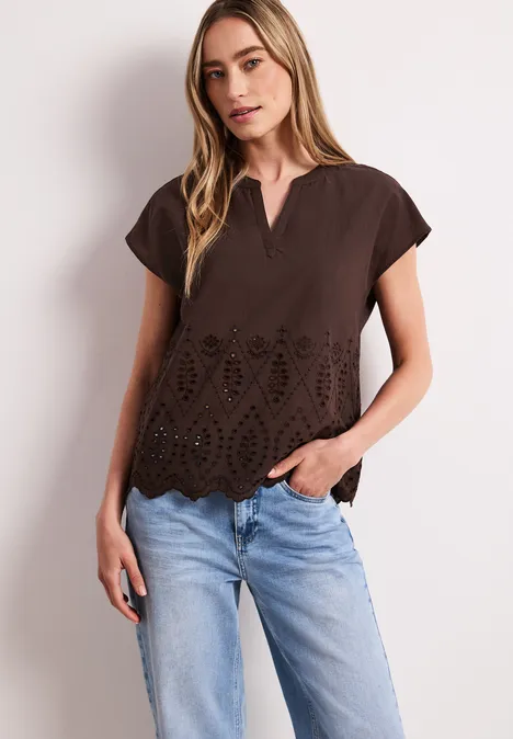 Vrouw draagt bruin kort mouwtop met V-hals en ajourdetails, gecombineerd met lichtblauwe jeans.