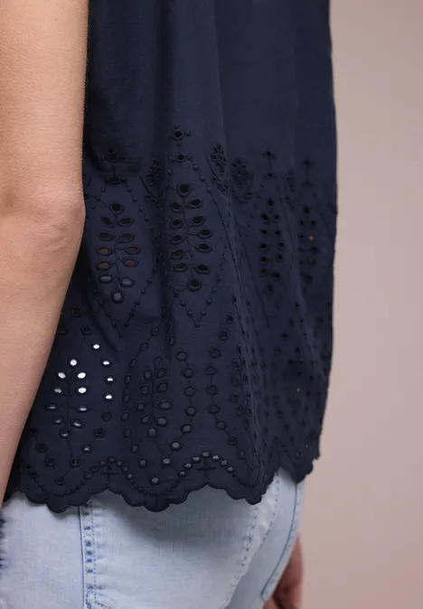 Persoon in lichte spijkerbroek en donkerblauw mouwloos topje met opengewerkt borduurwerk en gegolfde zoom.