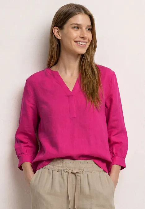 Frau mit pinker Bluse und beigen Stoffhosen steht lässig vor hellem Hintergrund.