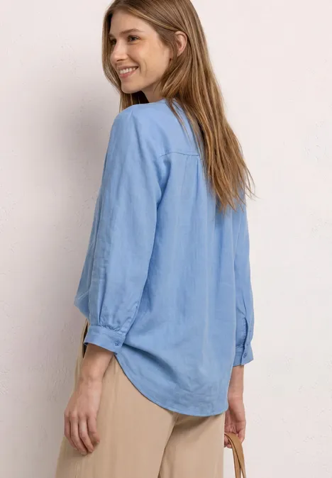 Seitliche Ansicht einer Frau mit hellblauem Hemd, beigen Hosen und kleiner Tasche.