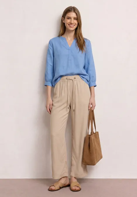 Frau trägt hellblaue Bluse, beige Paperbag-Hose, flache Sandalen und braune Wildledertasche.