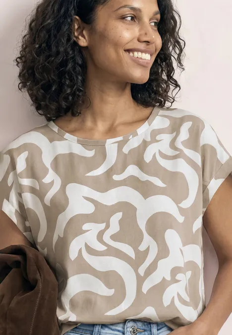 Eine Frau mit lockigem Haar trägt ein beigeweiß gemustertes Shirt und hält eine braune Jacke über dem Arm.