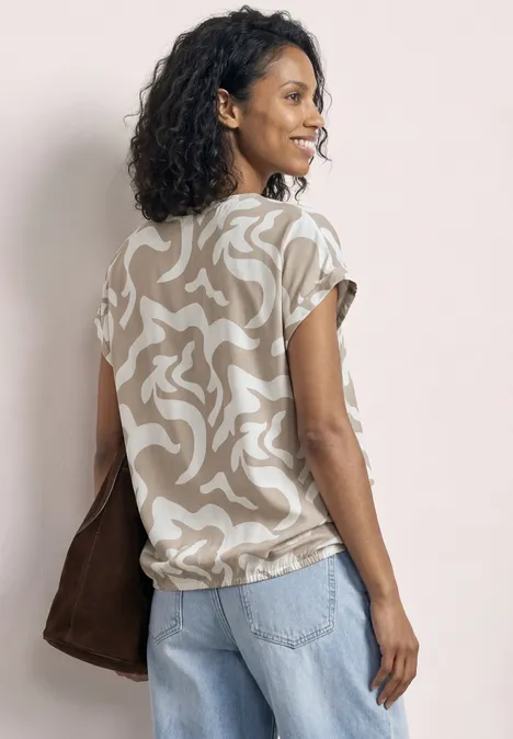 Eine Frau von hinten trägt ein beige-weiß gemustertes Shirt, hellblaue Jeans und eine braune Tasche.