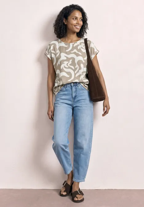 Eine Person trägt ein gemustertes T-Shirt, hellblaue Jeans mit Umschlag und schwarze Sandalen, dazu eine braune Tasche.