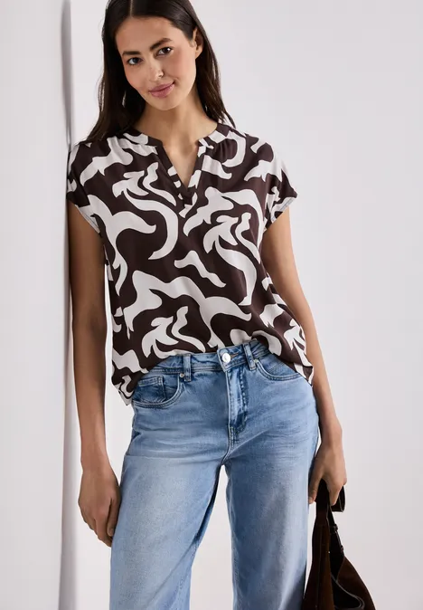 Frau mit schwarz-weiß gemusterter Bluse, Jeans und Tasche stehend vor hellem Hintergrund.