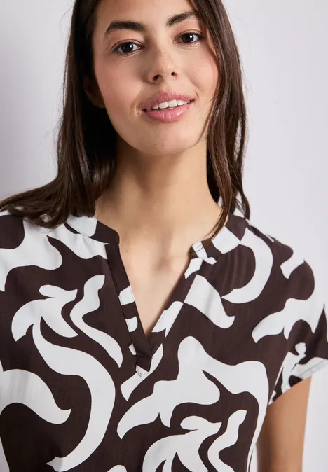 Frau mit braun-weiß gemustertem Kurzarmshirt und V-Ausschnitt vor neutralem Hintergrund.