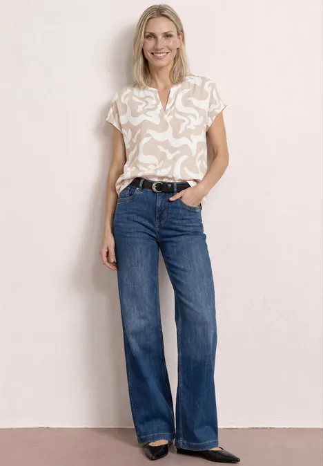 Frau trägt hell gemustertes T-Shirt, dunkle Schlagjeans und Gürtel, steht vor neutraler Wand.