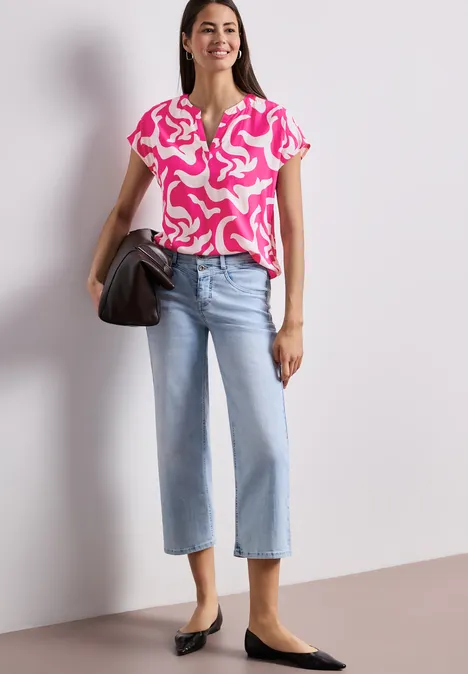 Frau in pink-weißem Muster-Top, weiten hellblauen Jeans, schwarzen Ballerinas und schwarzer Handtasche