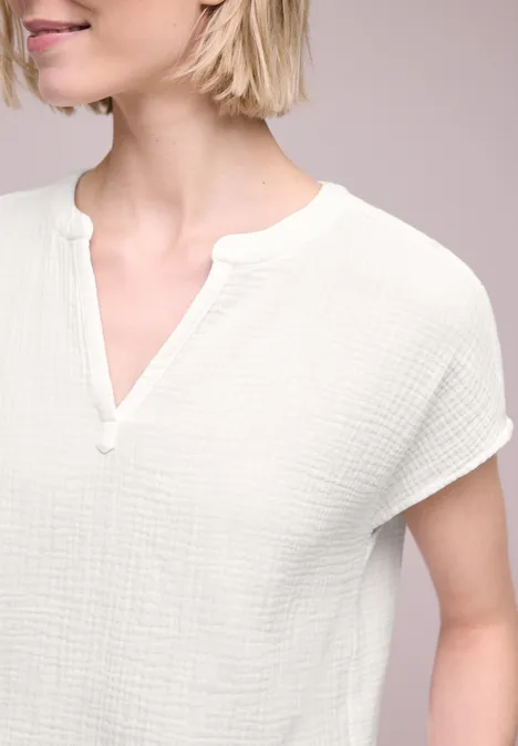 Eine Frau trägt ein weißes, strukturiertes Kurzarmshirt mit V-Ausschnitt vor neutralem Hintergrund.