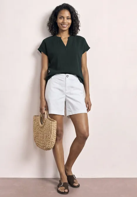 Model trägt schwarzes T-Shirt, weiße Shorts, schwarze Sandalen und Basttasche vor hellem Hintergrund.