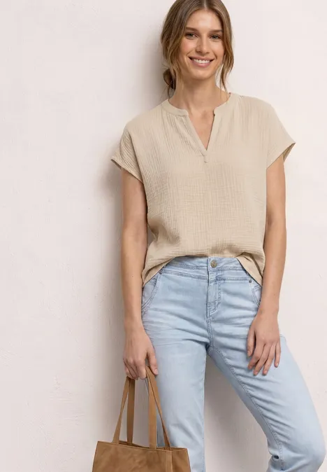 Eine Frau trägt ein helles, kurzärmeliges Oberteil, hellblaue Jeans und hält eine hellbraune Tasche in der Hand.