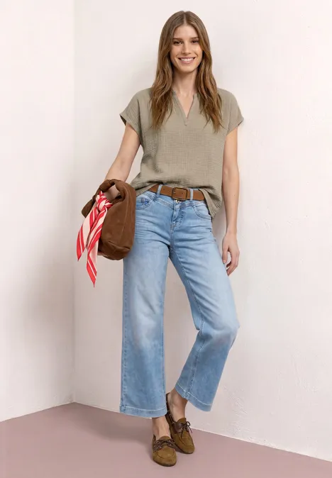 Eine Frau steht in der Ecke, trägt khakigrünes V-Shirt, hellblaue weite Jeans, braune Slipper, Tasche mit rotem Tuch.