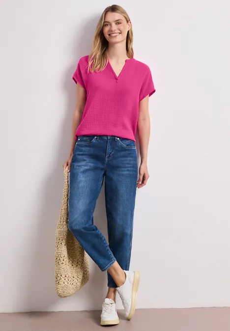 Frau trägt pinkes T-Shirt, Jeans, weiße Sneaker und hält eine Korbtasche vor hellem Hintergrund.