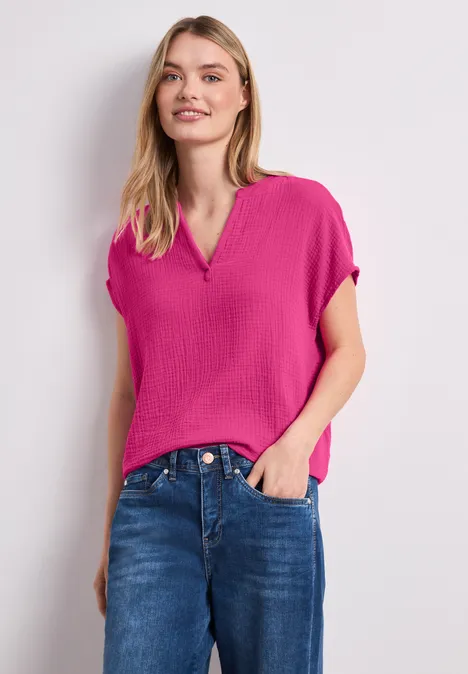 Frau in pinker kurzärmeliger Bluse mit V-Ausschnitt und blauen Jeans vor neutralem Hintergrund.