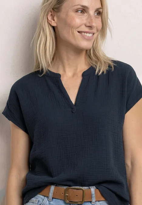 Frau mit blonden Haaren in lässiger dunkelblauer Bluse und braunem Gürtel, lächelnd vor hellem Hintergrund.