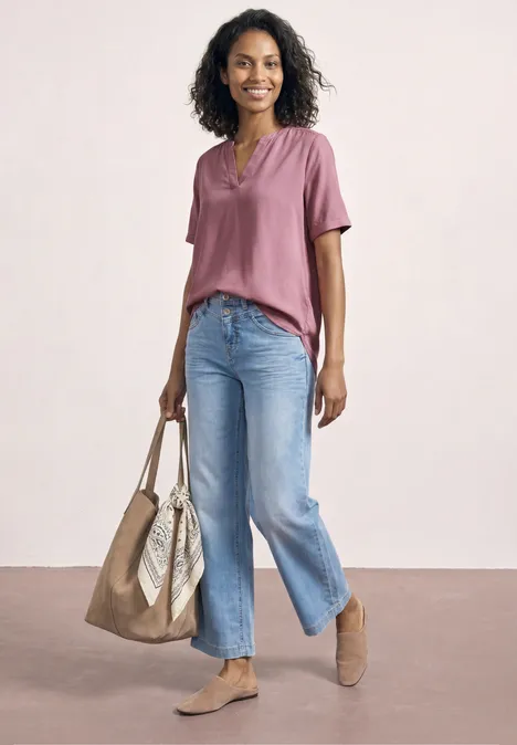 Frau mit lockigen Haaren in rosa Bluse, ausgestellten Jeans und flachen Schuhen, zwei Taschen.