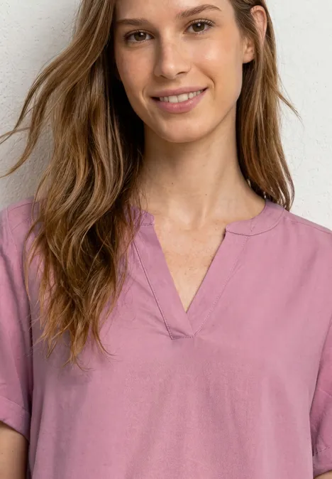 Frau mit verwischtem Gesicht, braunen welligen Haaren und rosa kurzärmeligem Oberteil vor neutralem Hintergrund.