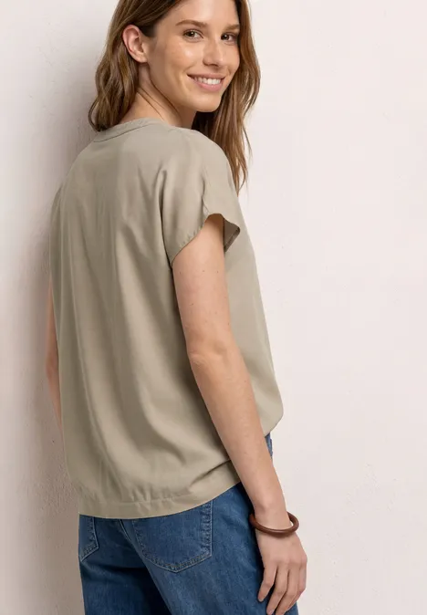 Frau von hinten in beigefarbener Bluse mit Kurzärmeln und blauen Jeans, Gesicht unkenntlich.