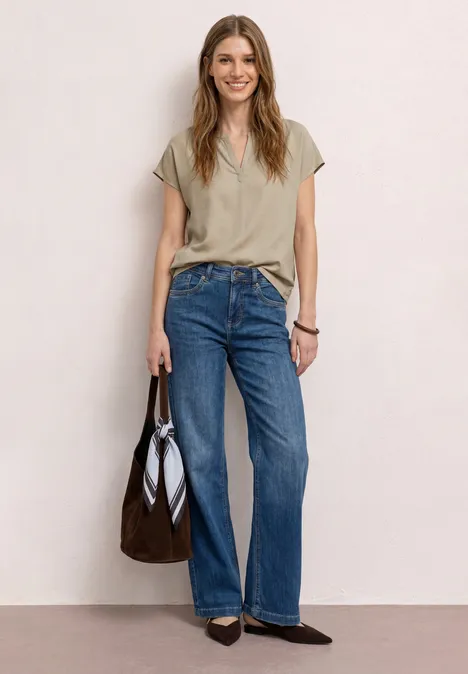 Frau vor hellem Hintergrund trägt beiges T-Shirt, blaue Jeans, schwarze Ballerinas und hält braune Tasche.