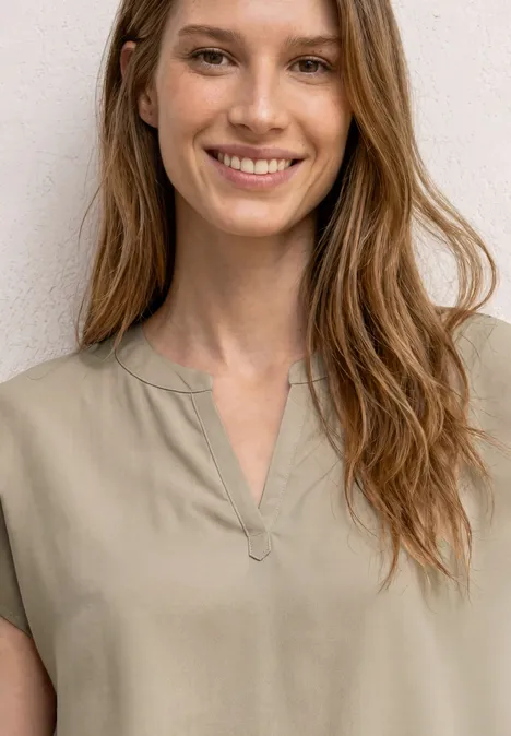 Eine Person mit langen, lockeren Haaren trägt ein beiges T-Shirt mit V-Ausschnitt vor einer weißen Wand.