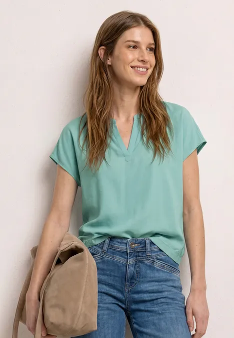 Femme portant un chemisier turquoise à manches courtes et un jean bleu, tenant un sac marron.