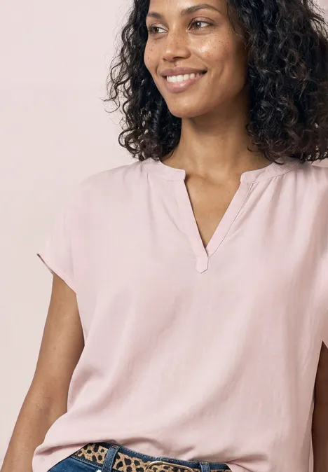 Oberkörper einer Person mit lockigem Haar, rosa Bluse und V-Ausschnitt vor hellem Hintergrund.