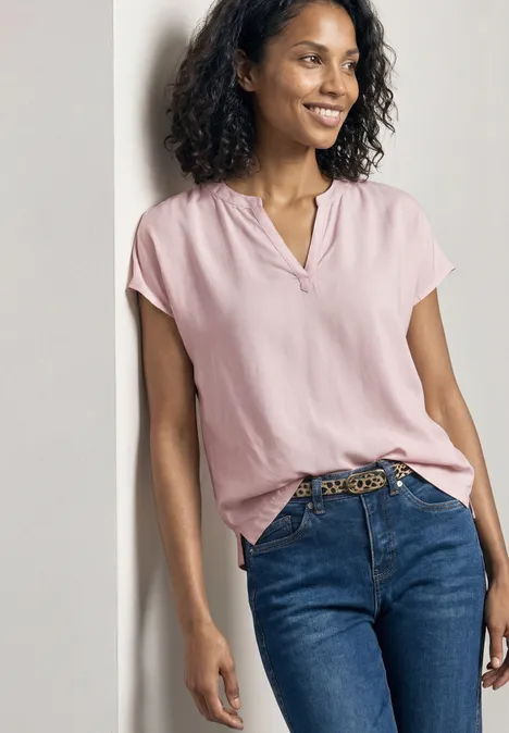 Frau in rosafarbener Bluse und Jeans lehnt lässig an einer Wand.